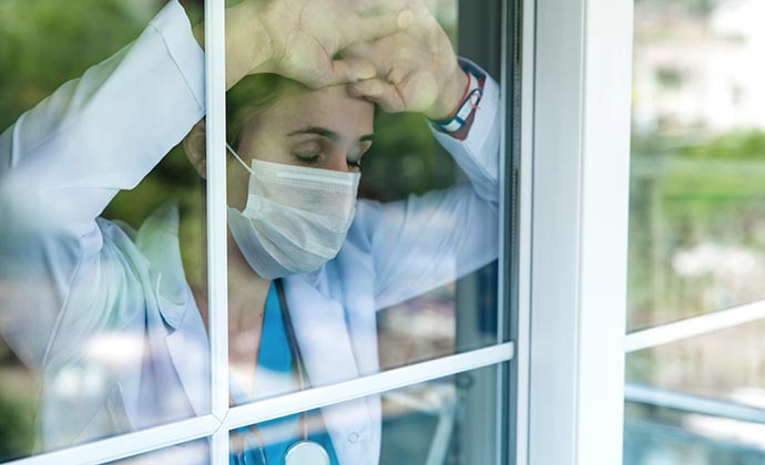 An exhausted healthcare worker, wearing PPE, looks out a window